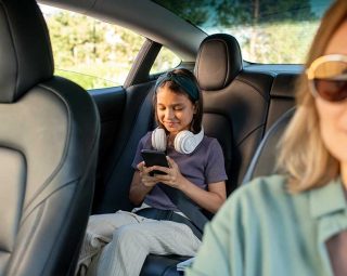 Happy schoolgirl with headphones scrolling in smartphone while sitting on backseat of car